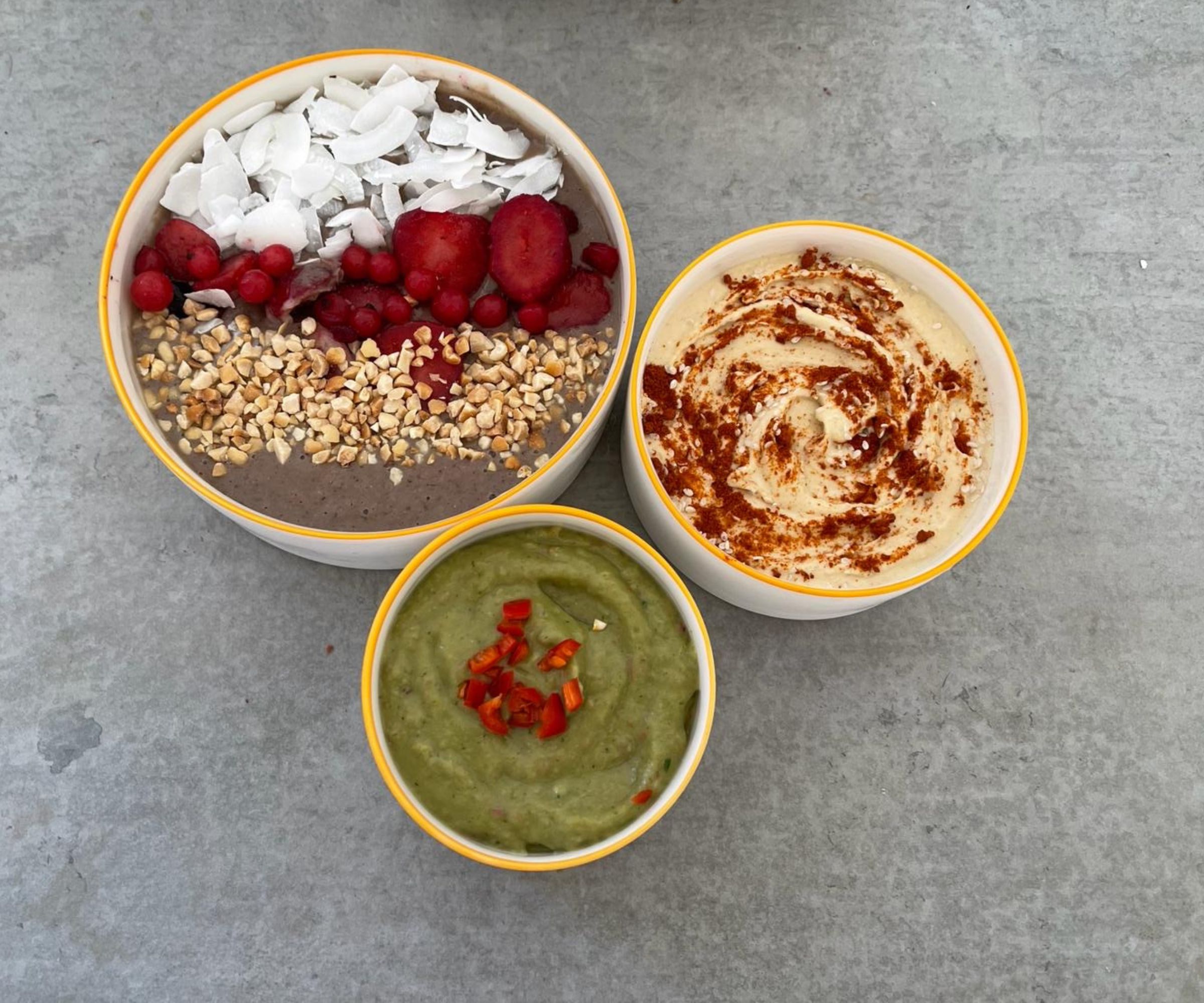 En smoothie bowl ved siden av guacamole og hummus på en grå benkeplate
