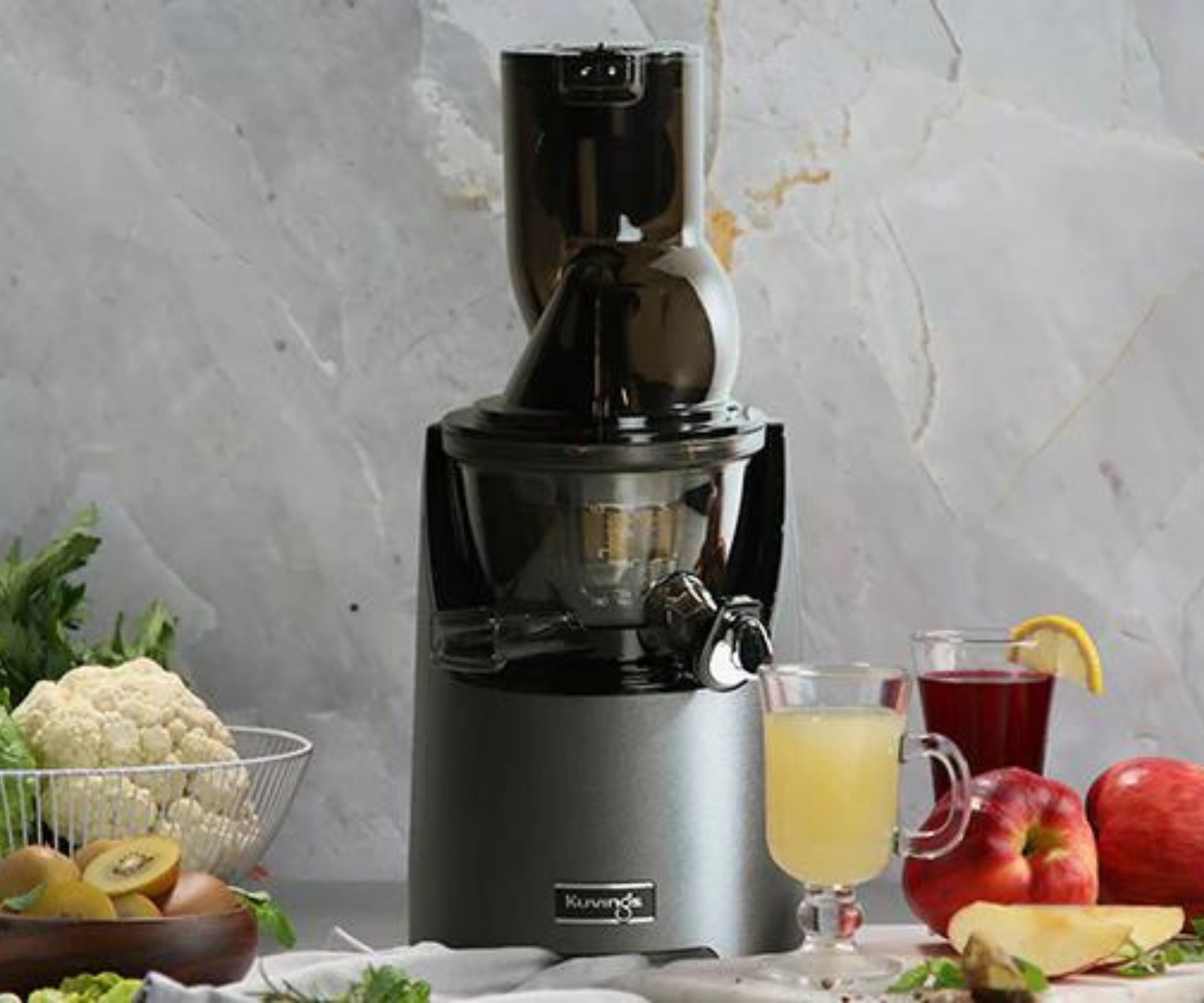 En Kuvings juicer mot grå bakgrunn, omgitt av frukt.