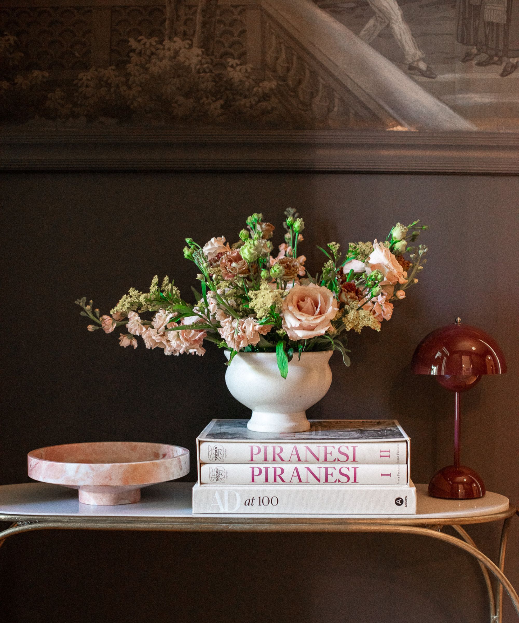 mørk brun vegg med konsollbord med vase med blomster, stabel med bøker, dekorativt brett og rød lampe