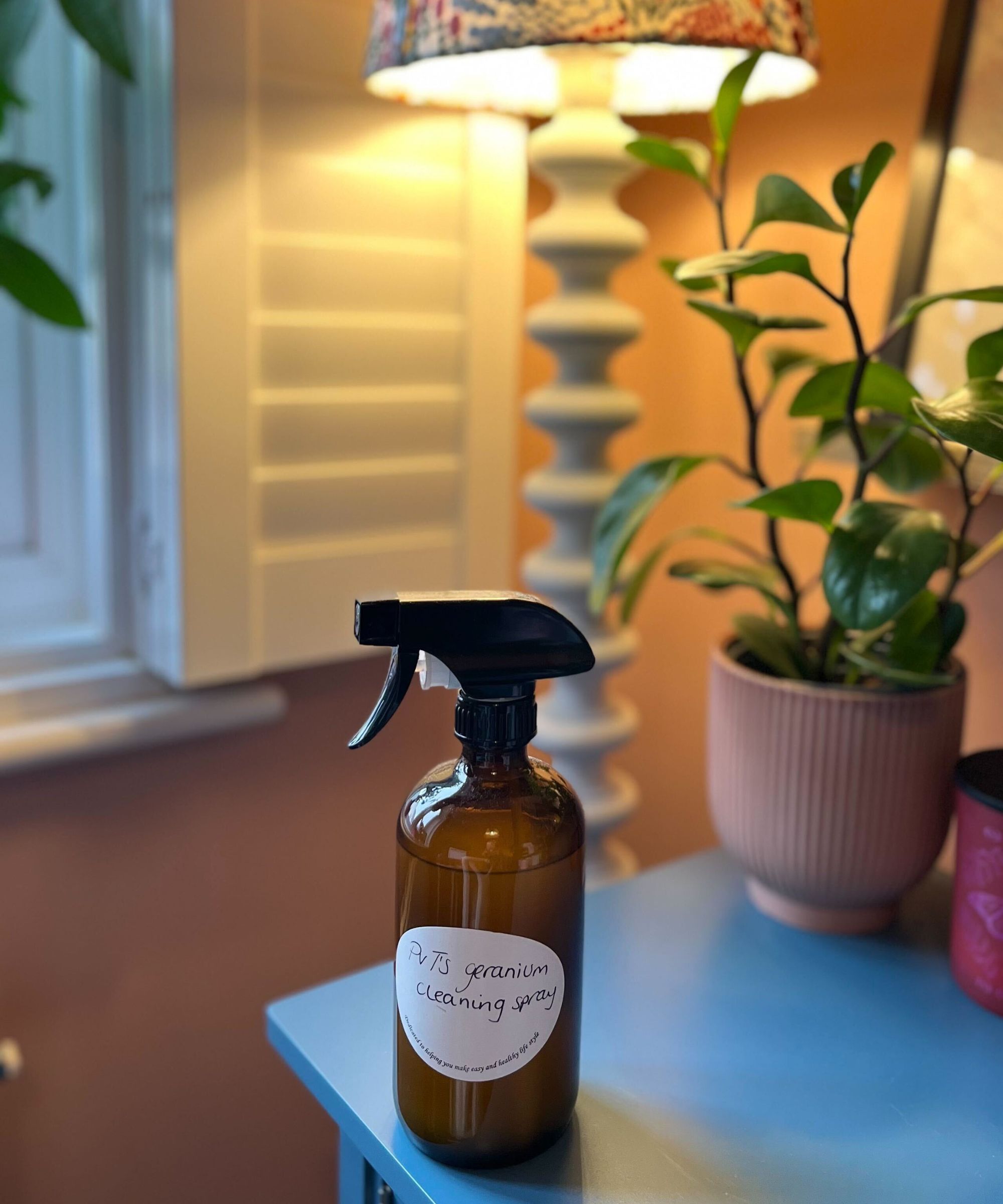 Min geranium-rensespray i en gul glassflaske med hvitt lokk og etikett på en blå skjenk mot en bakgrunn av terrakottamalt vegg med hvit bobbylampe og grønn plante i ribbet potte