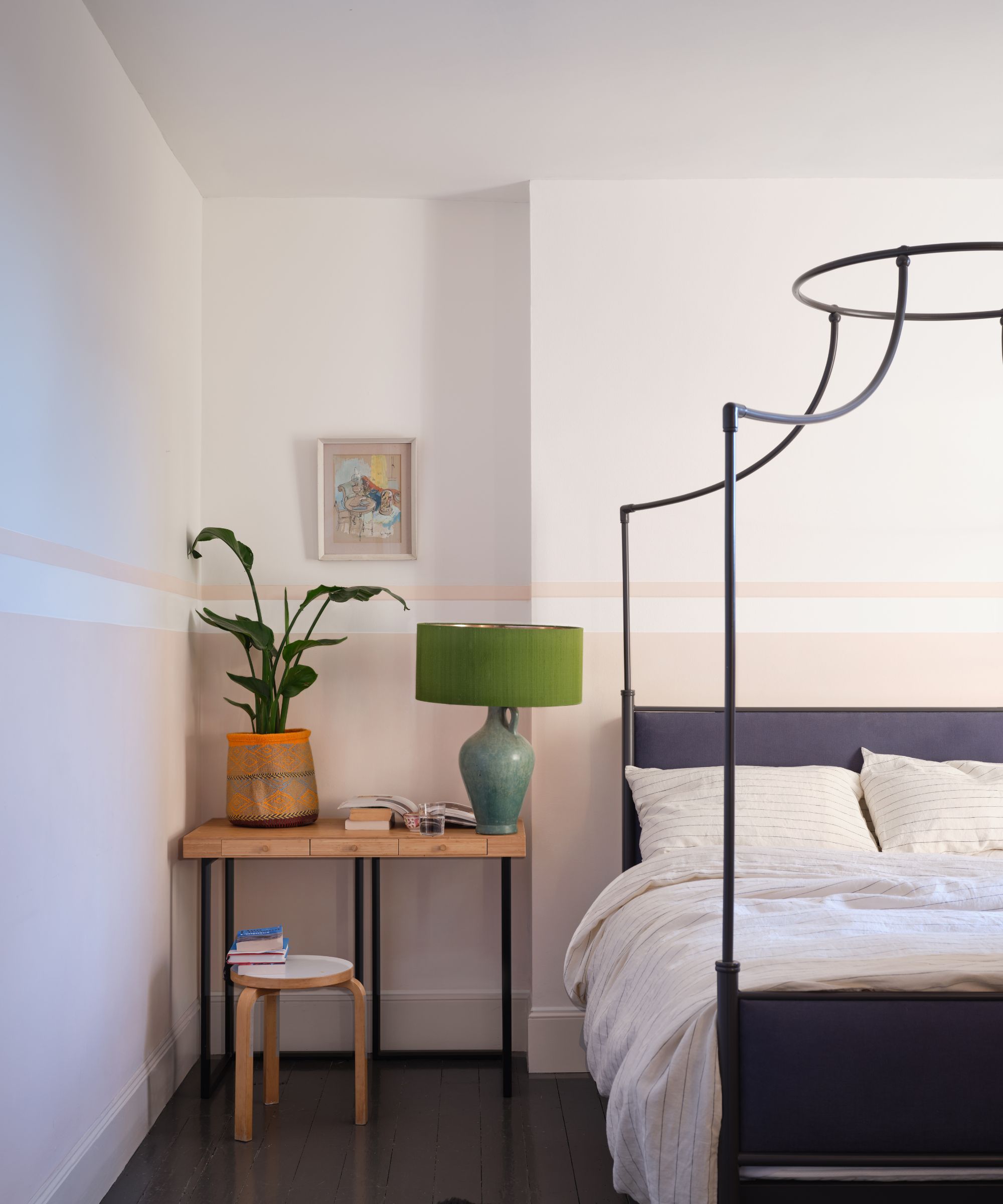 soverom med hvite vegger, blekrosa nedre halvdel av vegger, svart sengsramme, trebord i hjørnet med lampe og plante