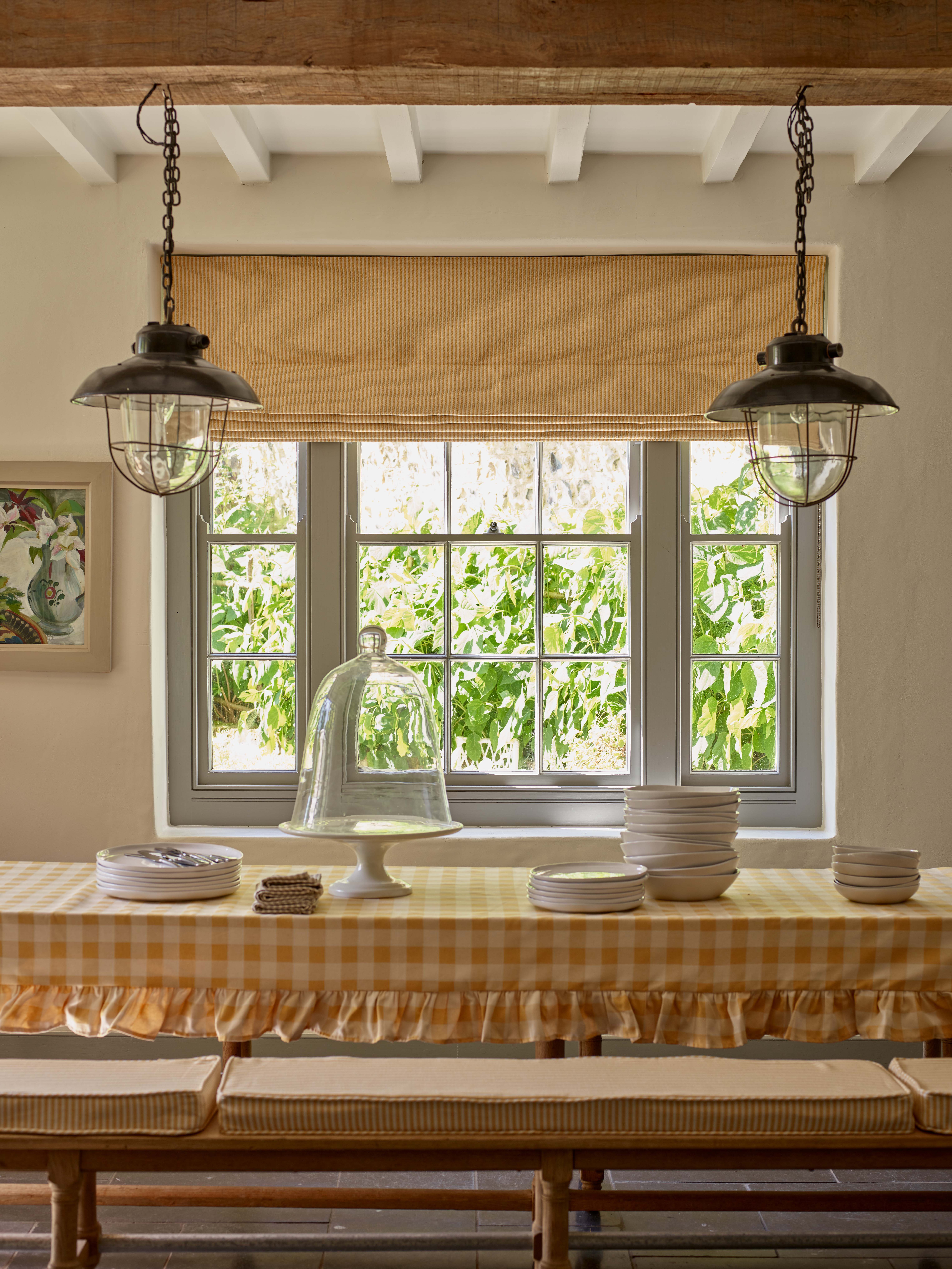 Farmhouse Spring Decor Idea With Yellow Gingham Table Cloth with Frills, Yellow Stripe Bench Sears og Matching Blind, Fisherman Style Pendant Lights, Beams, Artwork, Plates and Cake Stand on Table