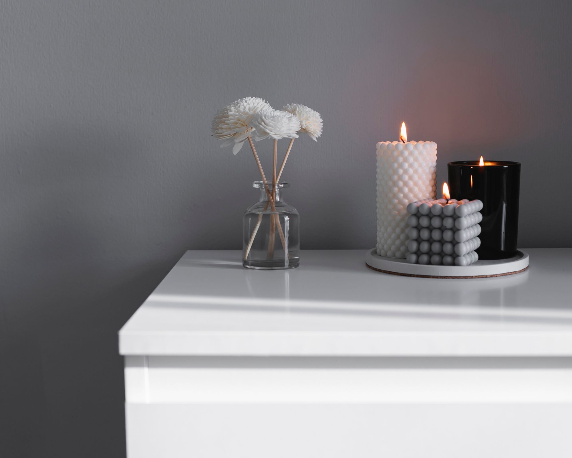 Tre lys som brenner på en hvit overflate ved siden av en liten vase med tre hvite blomster i mot en grå bakgrunn
