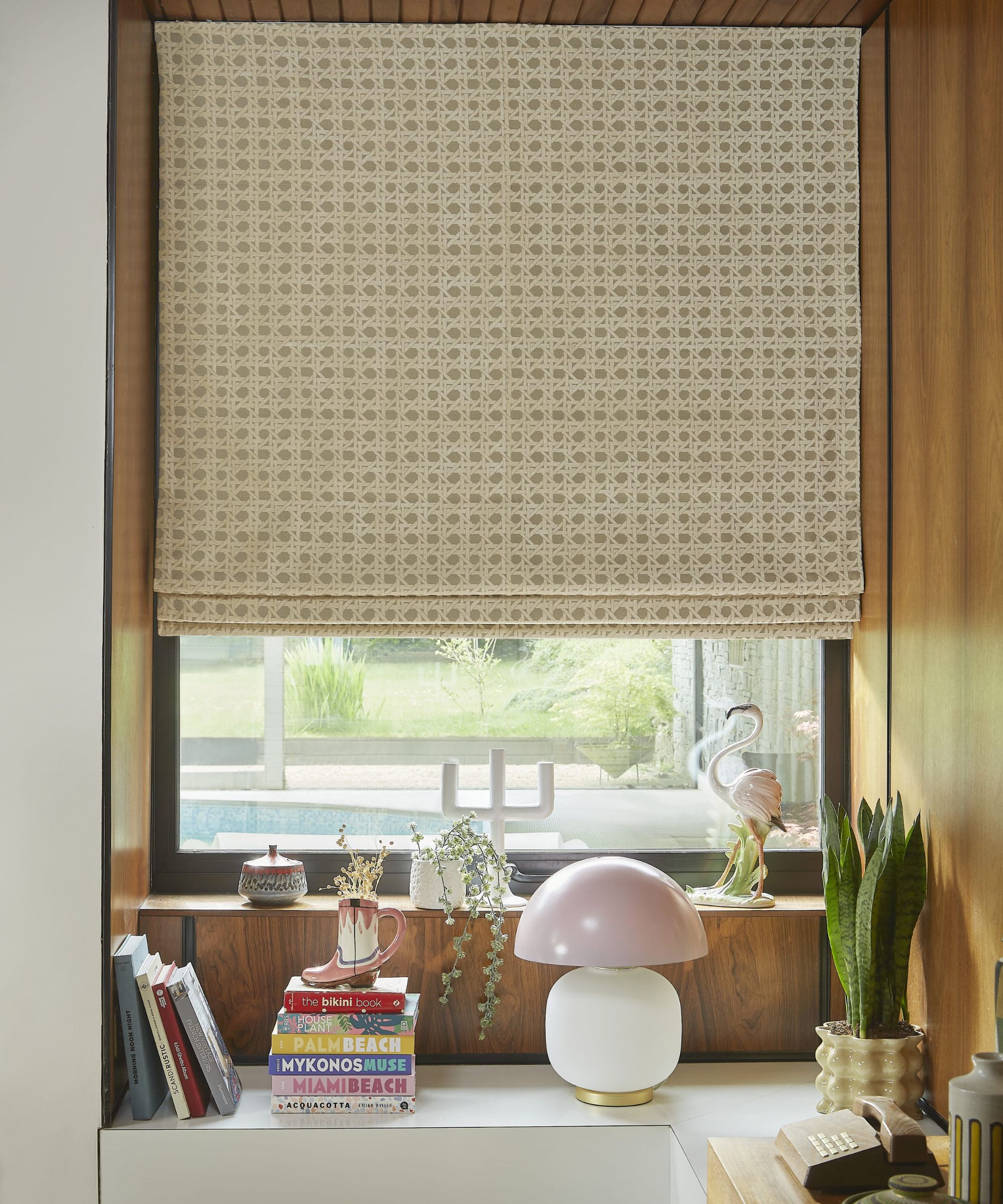 Et vindu med en brun og hvit blind, treveggpanel rundt det og fargerike bøker, en lampe og planter på hvithyllen under den