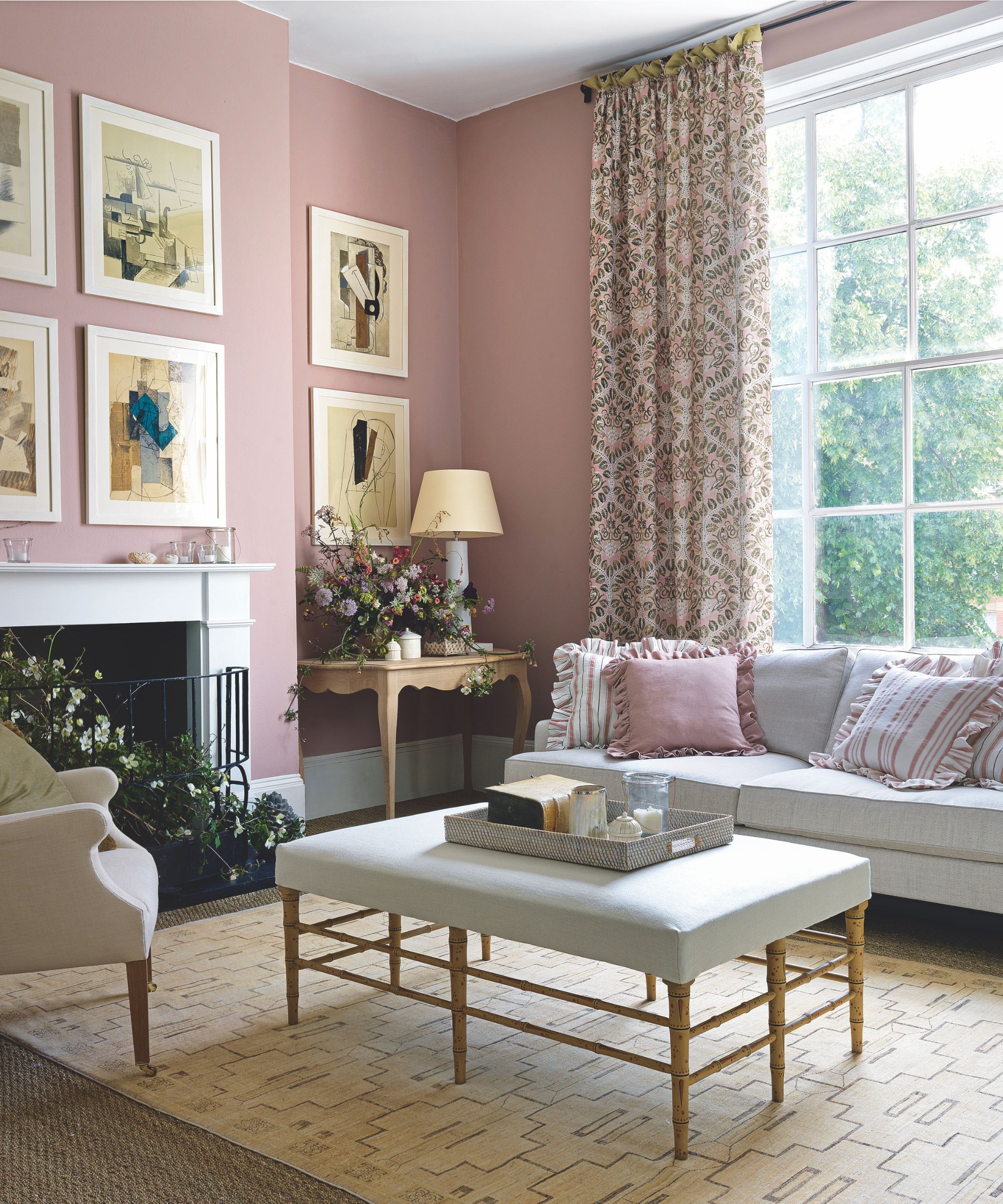 En rosa stue i vintage stil med mønstrede gardiner foran et stort vindu, med hvitt innrammede gallerivegg, en hvit mantelstykke, en grå sofa med frilly rosa og stripete puter med matchende fotstøtte med brett og en rødme rosa lenestol mot den