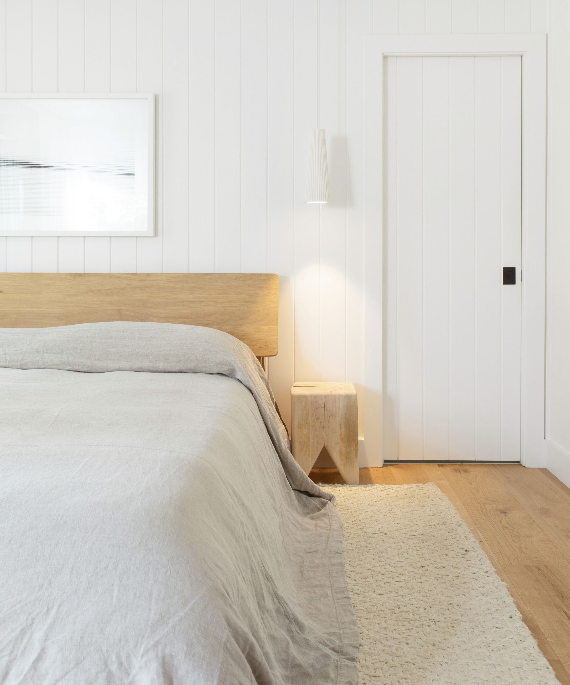 Nøytral skipslappet hvitt soverom med en tre -sengramme og hodegjerde, lite sidebord, ved siden av fotografisk veggkunst og grå lin sengetøy