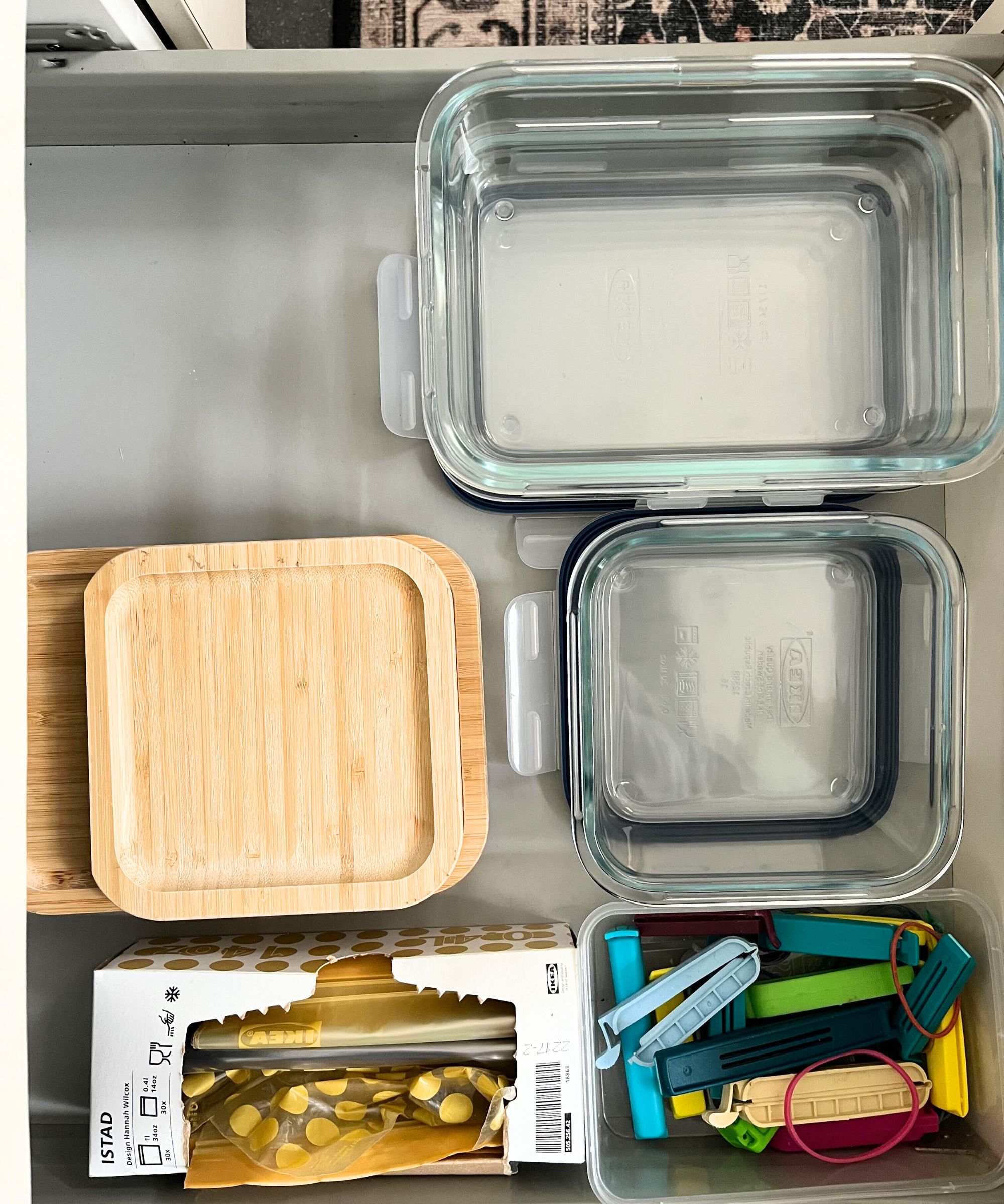 Rektangulære og firkantede glassmatbeholdere ved siden av bambuslokk, matposer av plast og matklipp i plast i en kjøkkenskuff.