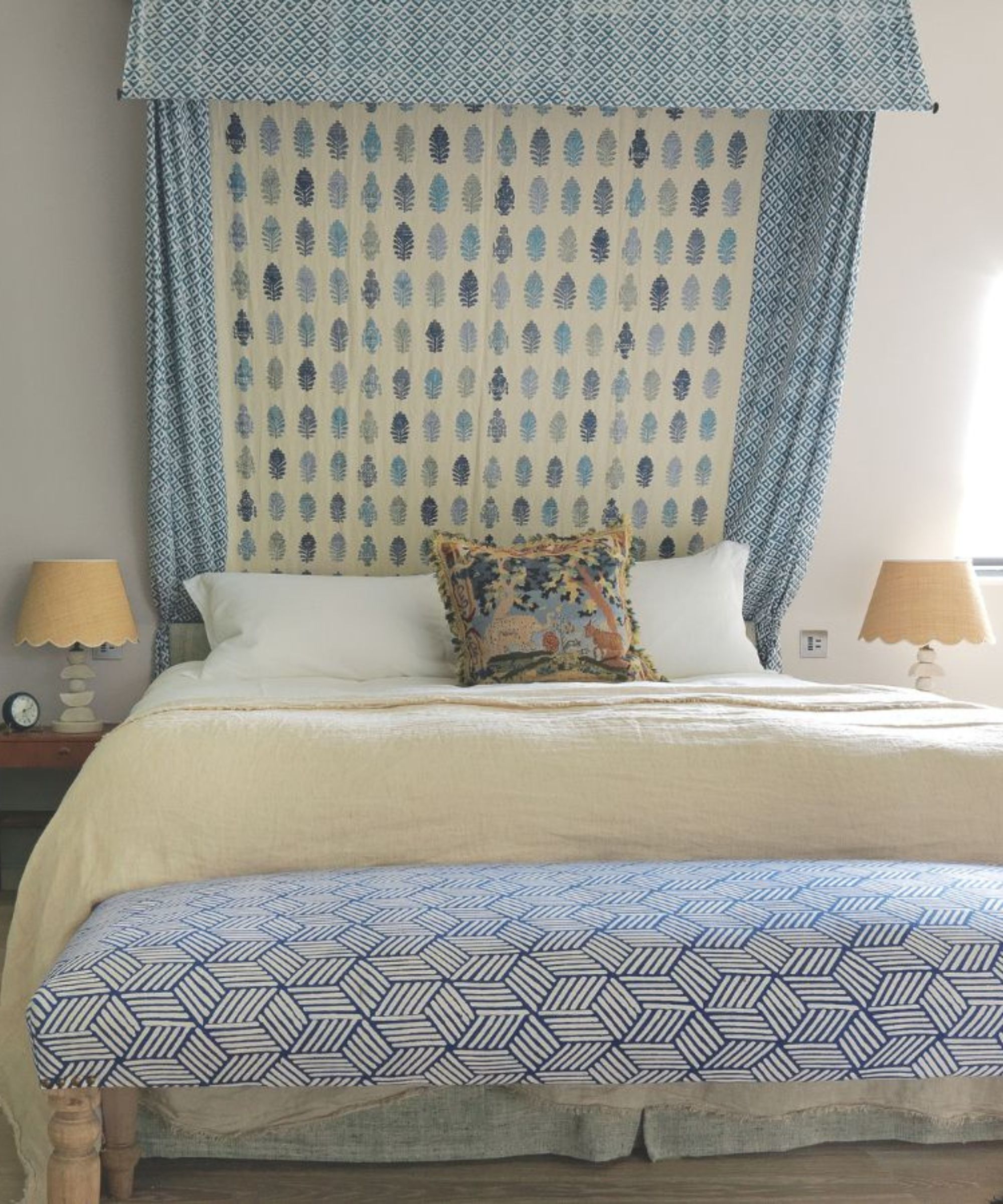 Et blått soverom med en mønstret seng kalesje og ottomansk, matchende beige kamskjell lamper hver side av sengen med hvite puter, beige teppe og mønstret pute