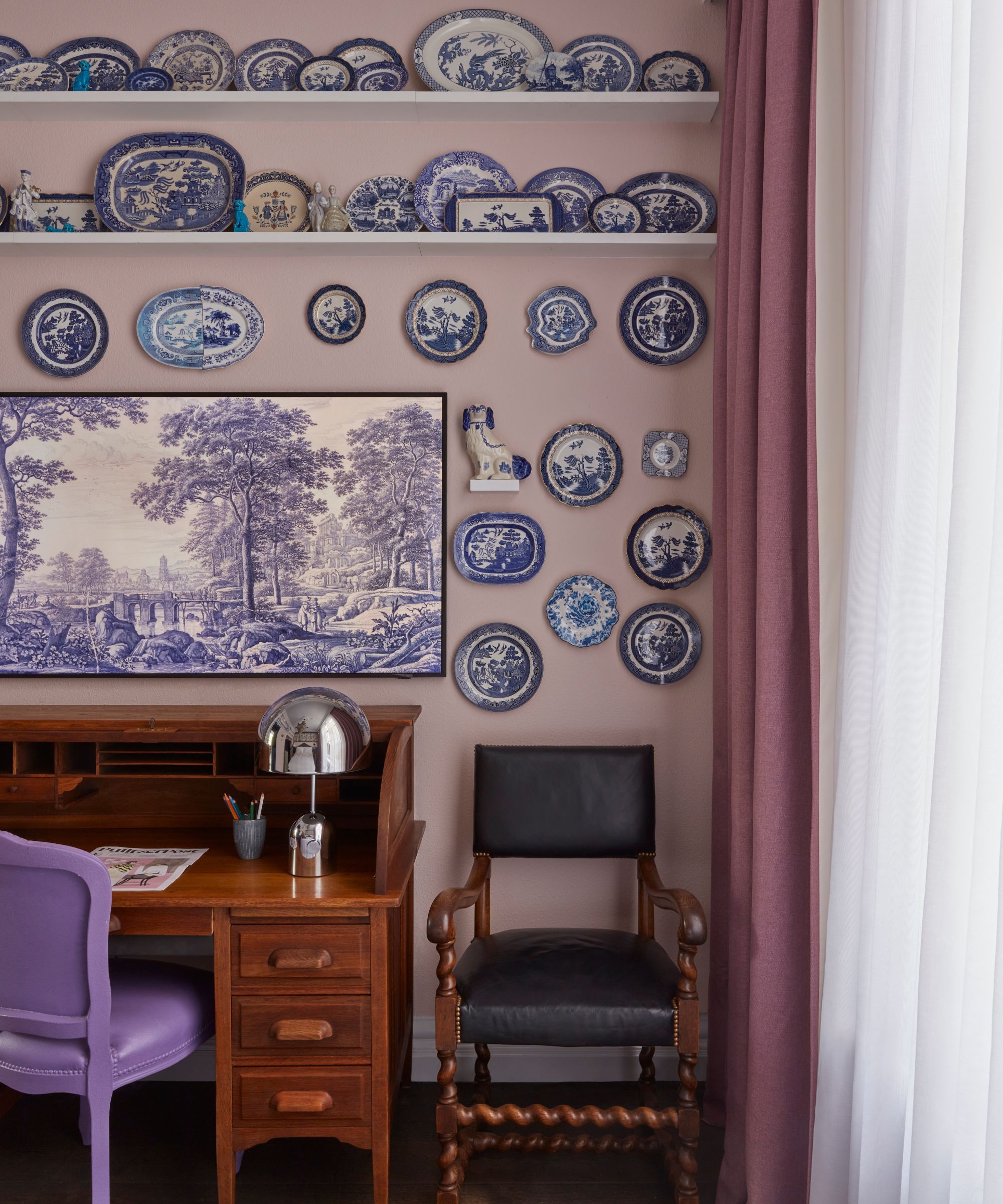 Lilla rosa hotell soverom med en stor samling av blå og hvite porselen veggplater og en ramme -TV hang over et tre -vintage skrivebord med en syrinstol og moderne sølvbordlampe