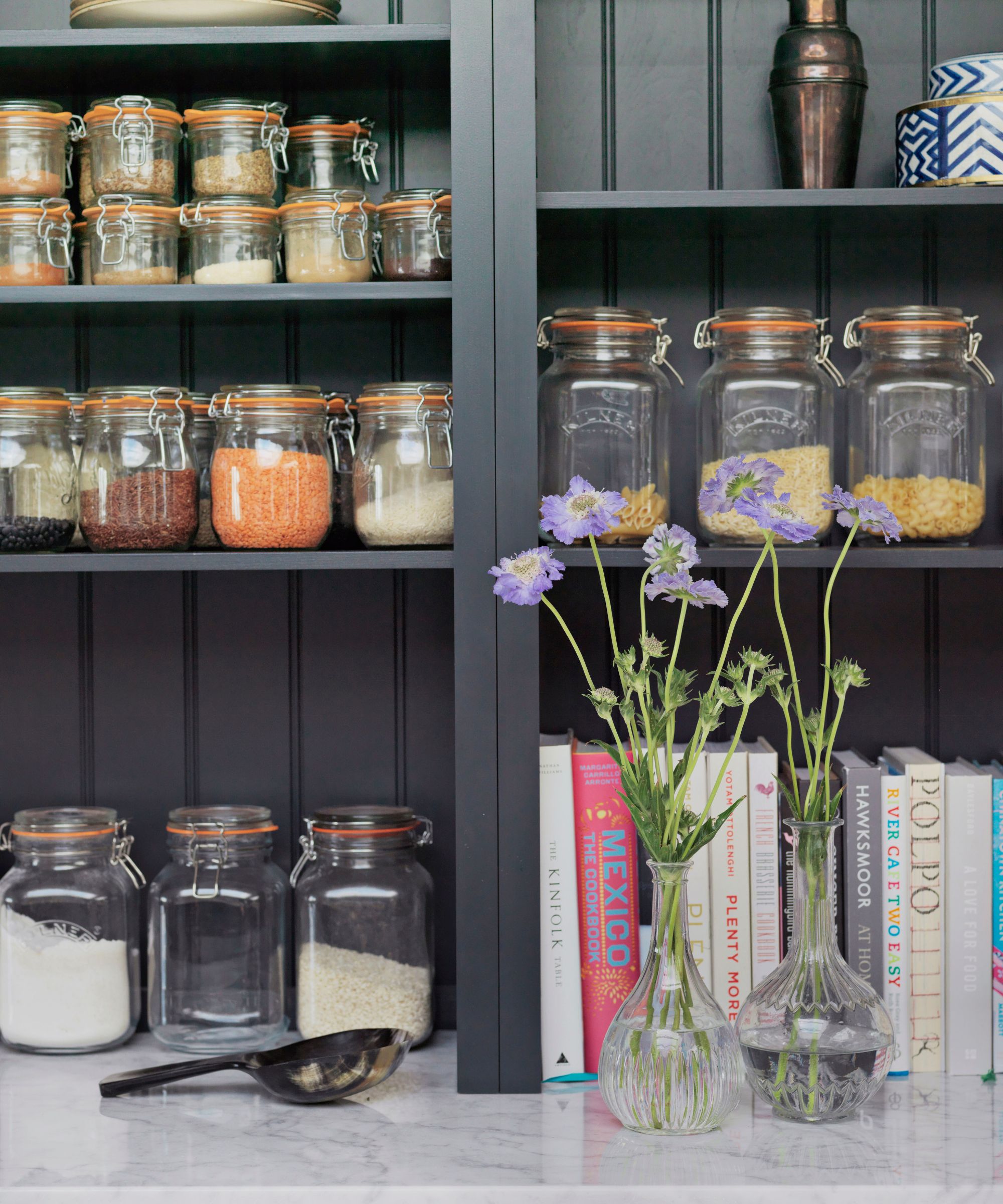 Mørkegrå spiskammer med Kilner -krukker, oppskriftsbøker og en kobbercocktail shaker, med glassvaser fylt med lilla blomster