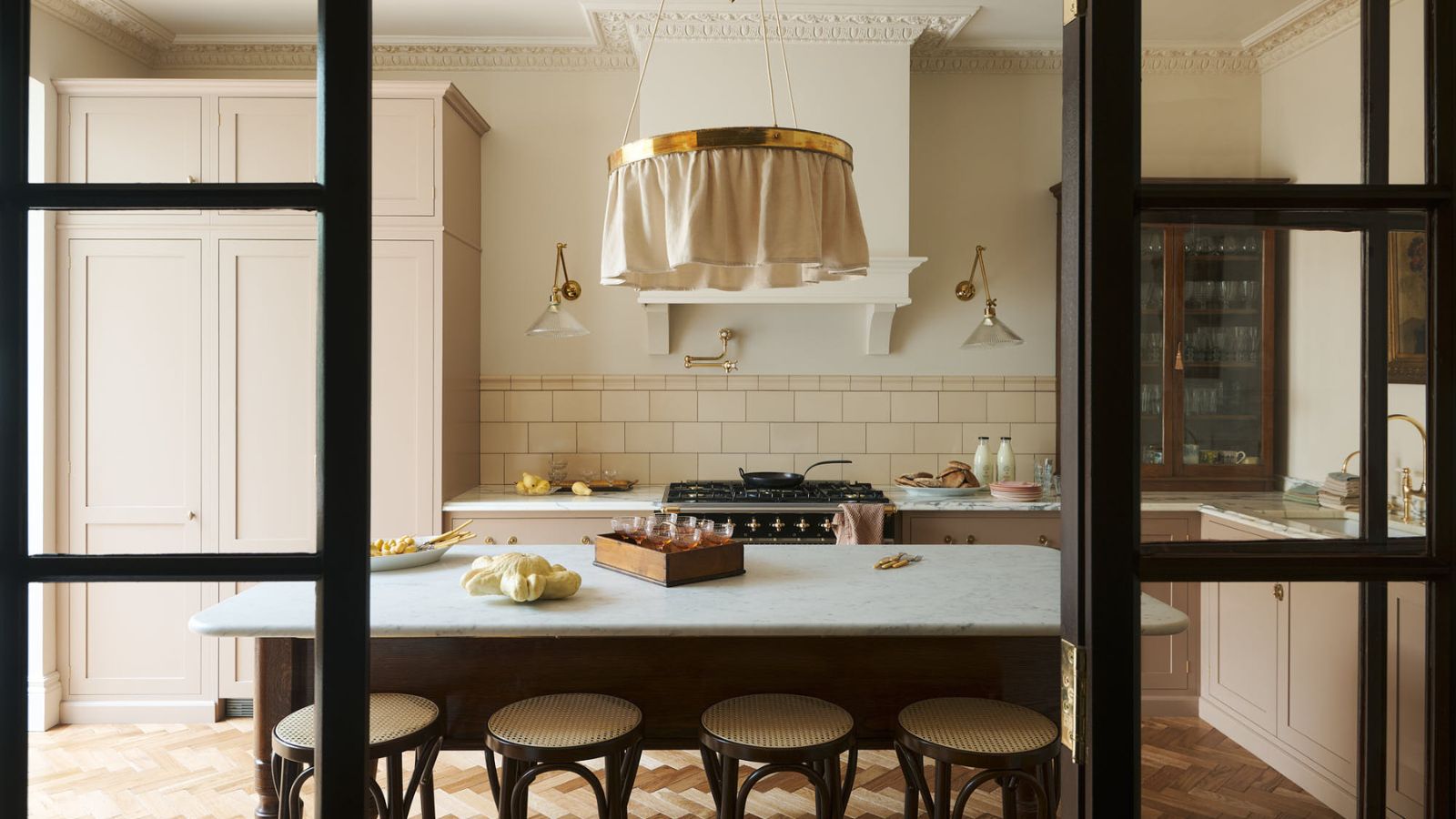 Character peachy pink kitchen viewed through internal black Crittal doors. There is a marble island table with wooden backless bar stools in view. In the background, there is a splashback tiled stove area and built-in pink, and glass front brown cabinets.