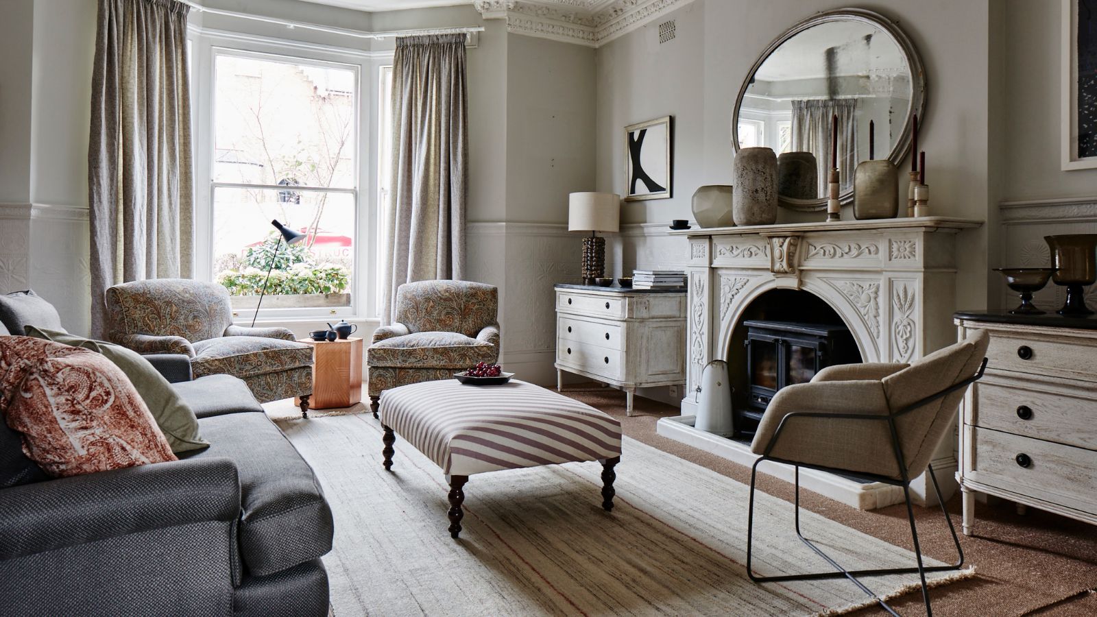 Neutral living room with large white mantelpiece, circular mirror, white rug, and grey sofa. In the centre is a striped footrest. There are large bay windows looking over a street behind in the back.