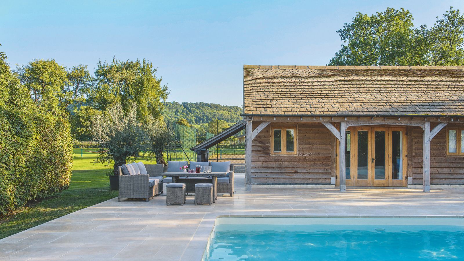 Large very clean and well-maintained outdoor paved area, showing pool house, outdoor dining furniture, and the edge of a pool in foreground
