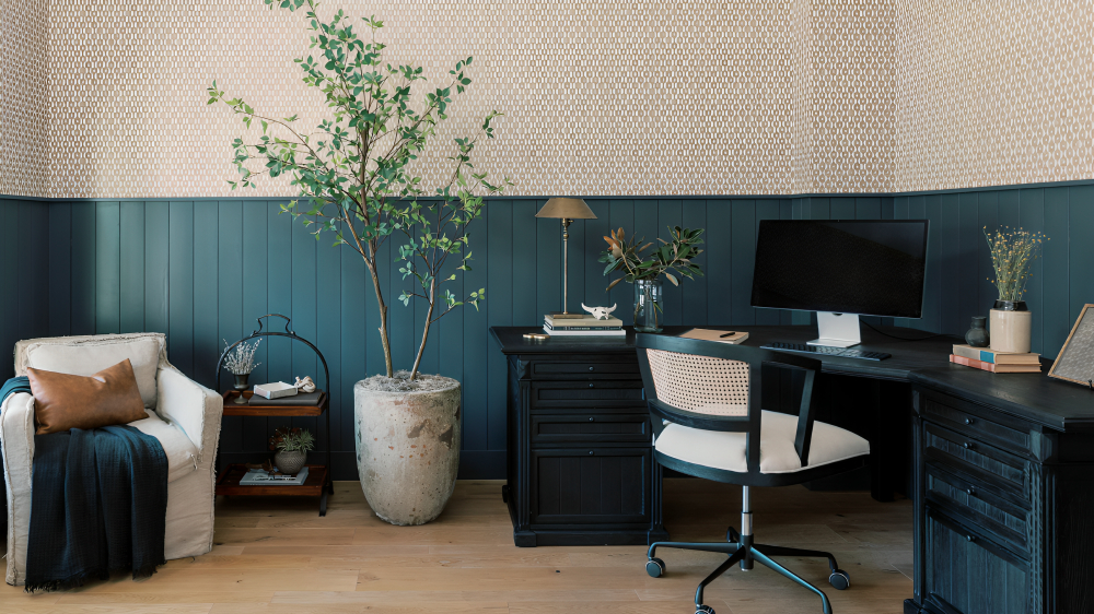 A home office with blue wall panelling on the bottom half of the wall, with a black corner desk, and a white armchair in the opposite corner.