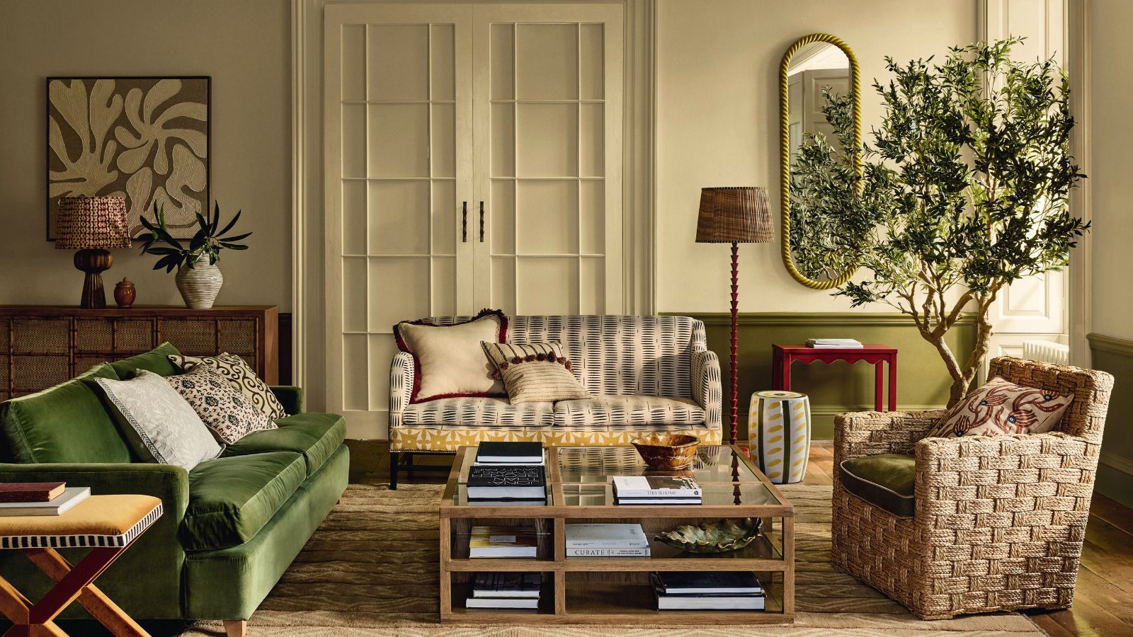 A warm toned living room with a green sofa, white patterned sofa, and a wicker chair. An olive tree in the background, and a glass square coffee table.