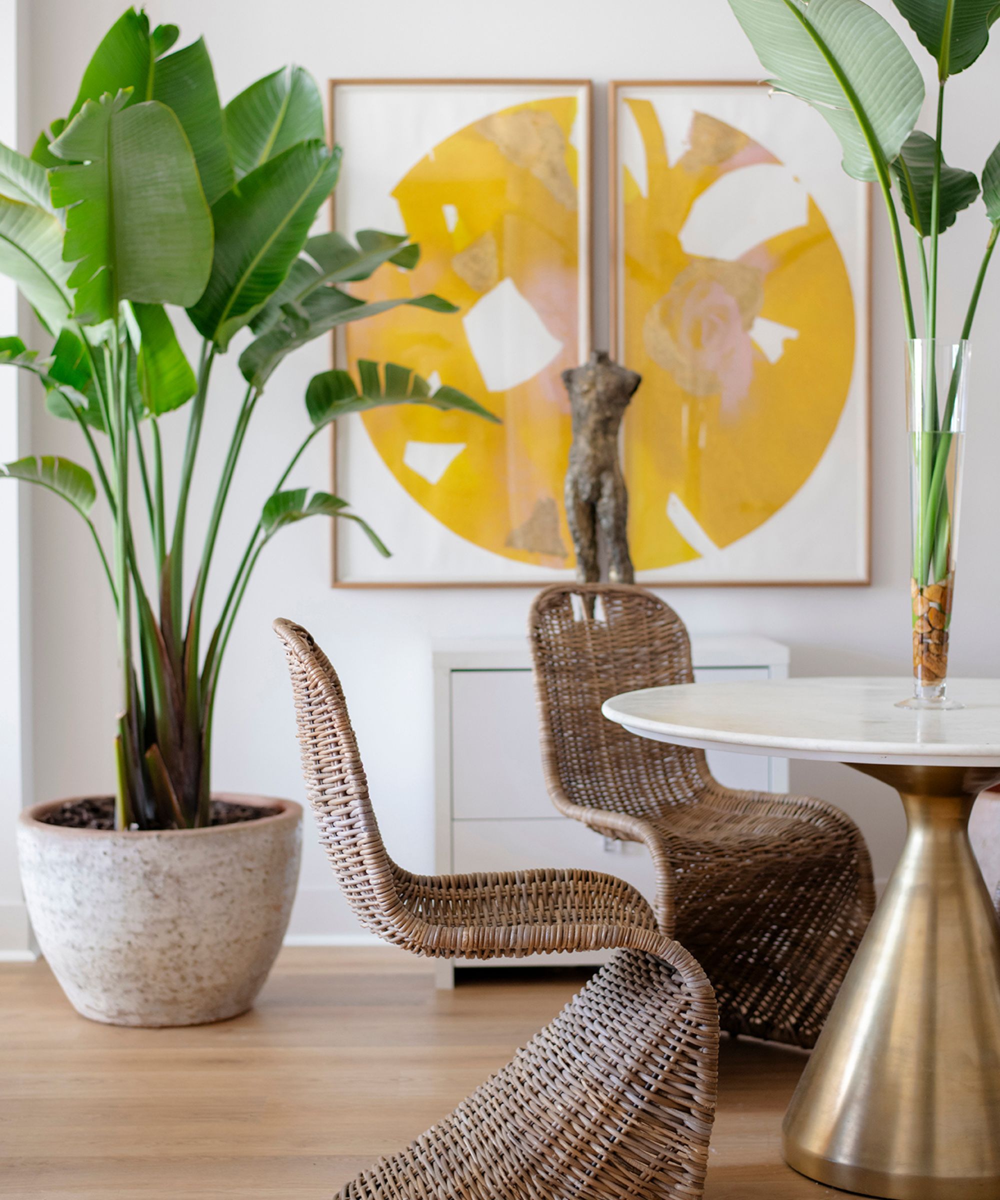 Levende spisestue med to rammer som ligner en sirkulær, abstrakt, gul design, en stor plante i keramisk vase, to buede kurvstoler og elegant bord med blomster i vase, på tregulv.