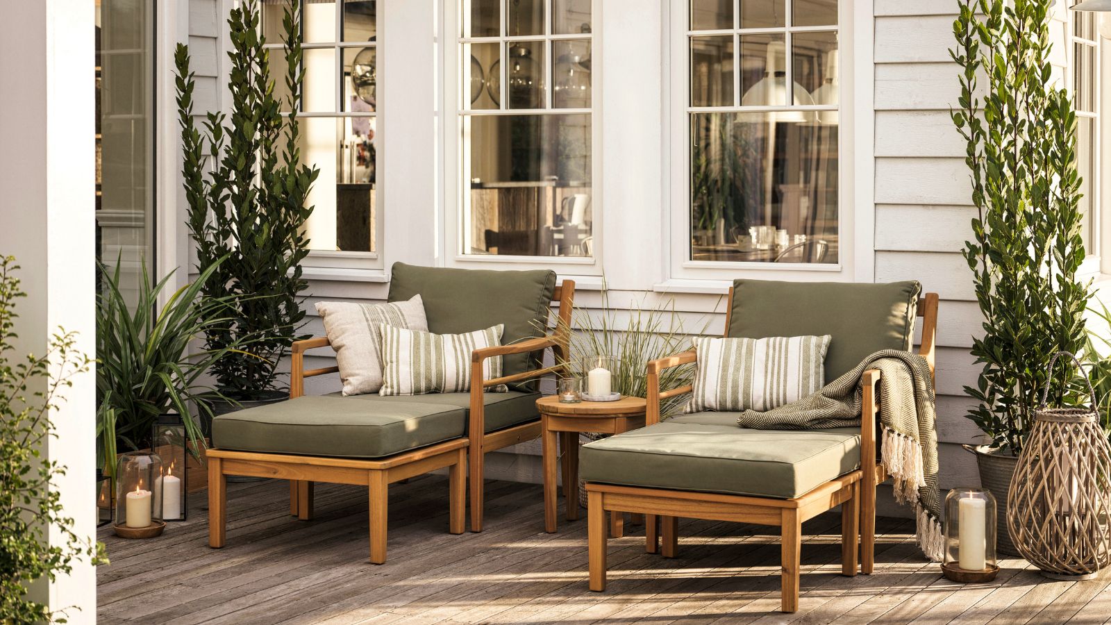 Two wooden sun loungers on a wooden patio, with green cushions. In front of a white sided house.
