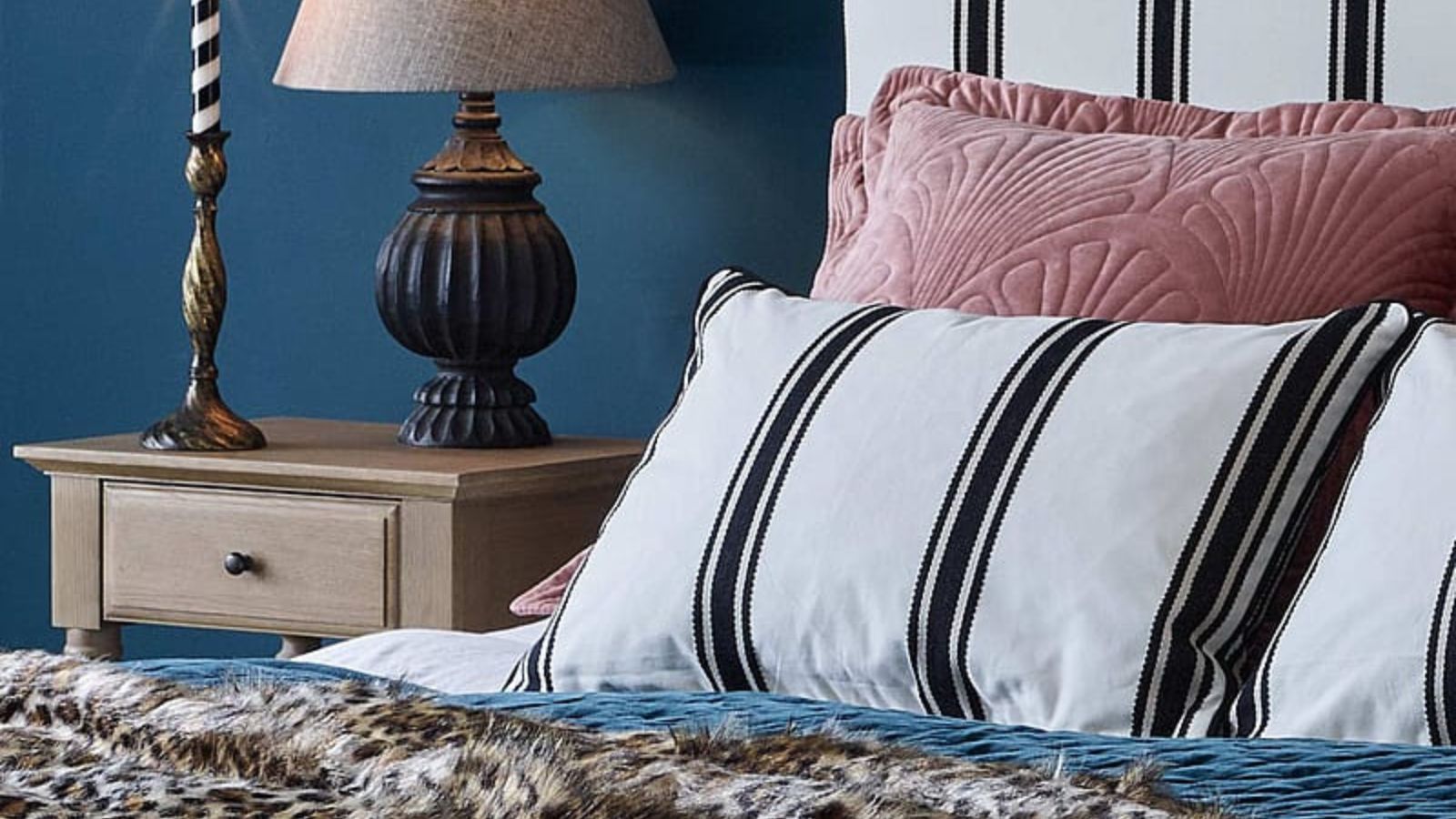 A bed with a white and black stiped throw pillow on top of a pink throw pillow, against a blue wall. A small wooden nightstand with a lamp and a candlestick beside it.