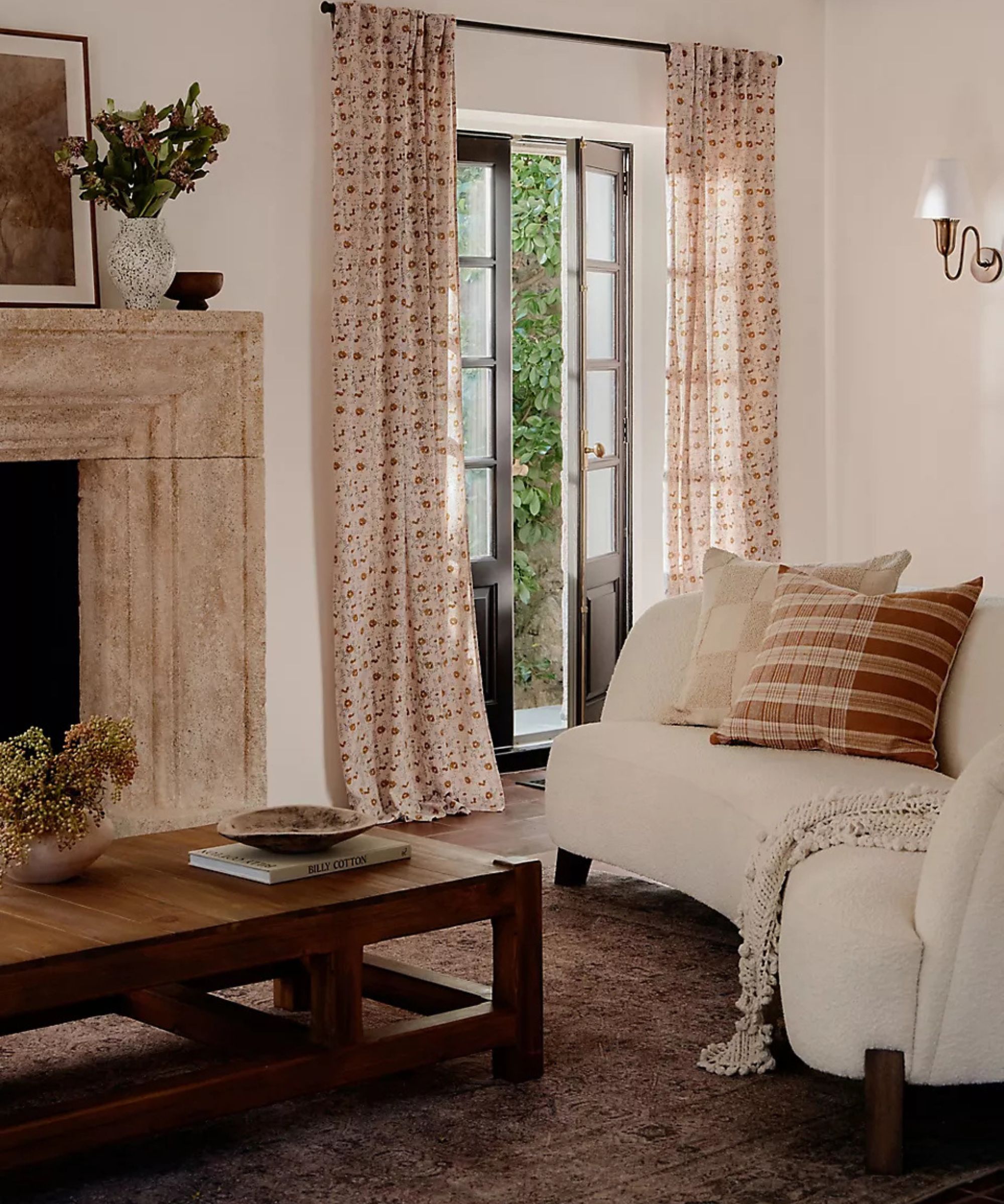 Moderne rustikk tidløs stue designet av Amber Lewis for Anthropologie med en steinpeis, mørk vintage-stil, buet moderne boucle sofa og blomsterutskrift gardiner hang på franske dører