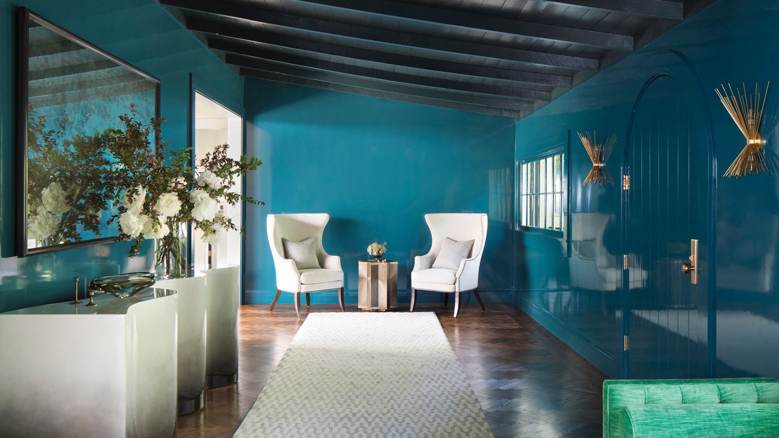 Room with sleek blue walls, two white armchairs in background, a white rug runner, marble-esque shelf with ornaments and flowers to the left.
