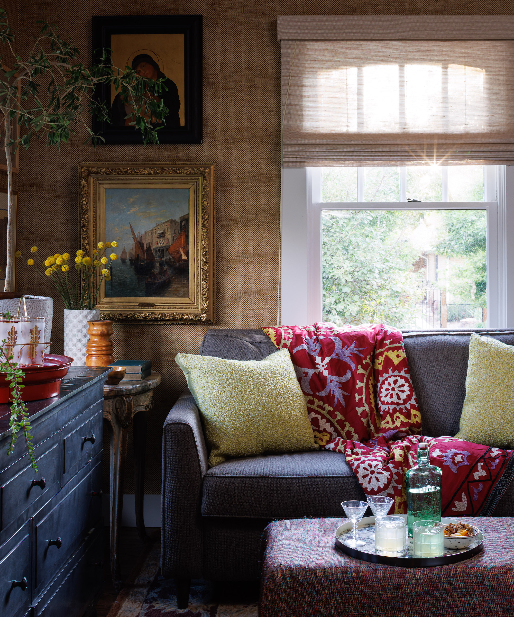 Stuen med sofa med grønne puter og drapert rødt mønstret kast, ottomansk med brett og barware, skuffer og veggkunst